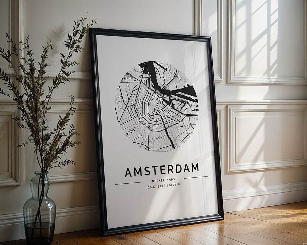 The FN Prints Amsterdam City Map, a minimalist black circular street and waterway design, leans elegantly against a white paneled wall. A tall plant in a clear vase on the wooden floor adds Scandinavian flair as light streams from the right.