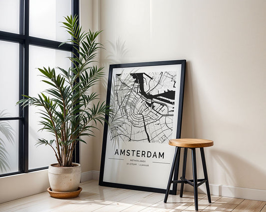 An Amsterdam City Map by FN Prints in a black frame leans against a white wall, epitomizing Scandinavian style. A potted plant sits left, and on the right is a sleek wooden stool. Sunlight filters through the window, casting elegant shadows on the floor.