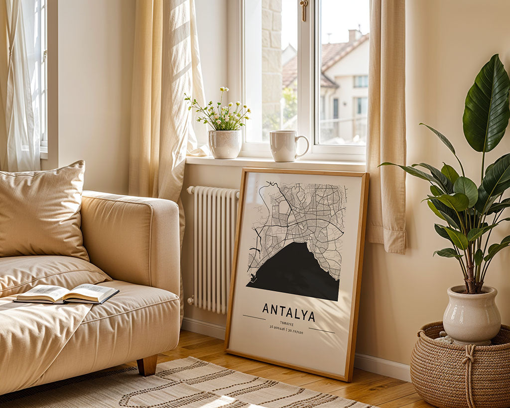 A cozy living room features a beige sofa with a book, potted plant, and FN Prints' Antalya City Map on fine art paper. Sunlight highlights the map beside a mug and flower pot on the windowsill, while neutral decor and a woven rug complete the scene.