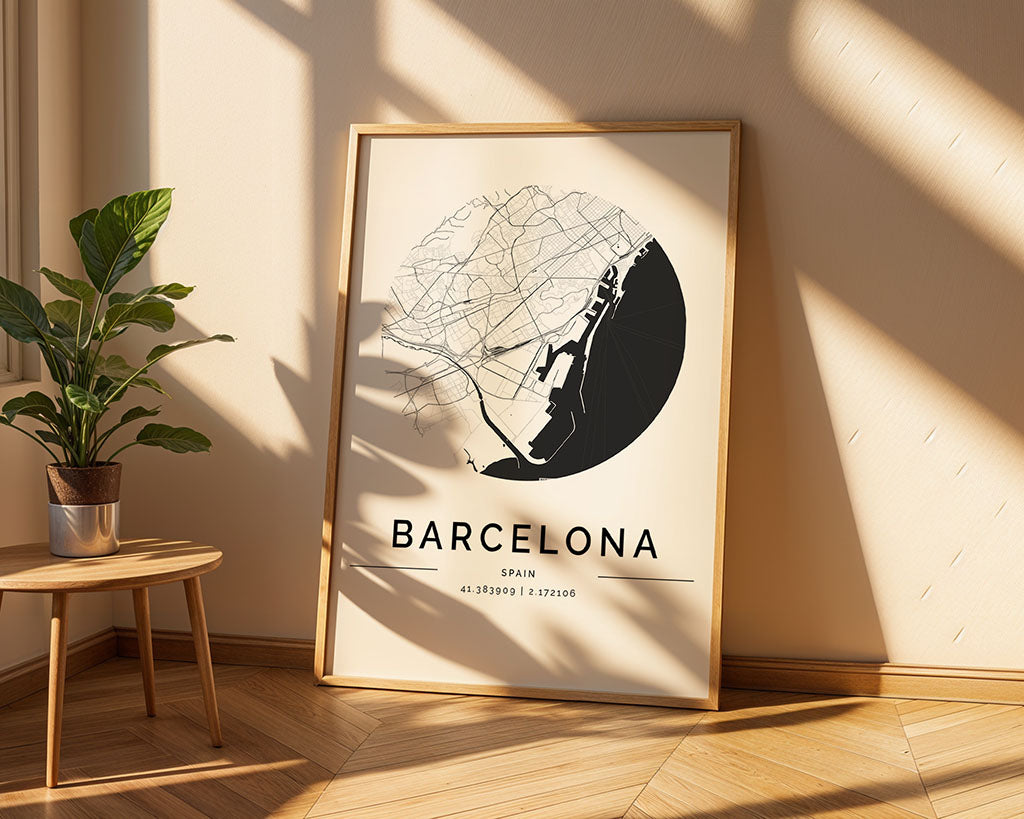 A FN Prints Barcelona City Map poster leans against a light-colored wall. The black city streets on a circular white background are ideal for urban design fans. A small potted plant sits nearby on the wooden floor, as sunlight casts shadows across the scene.