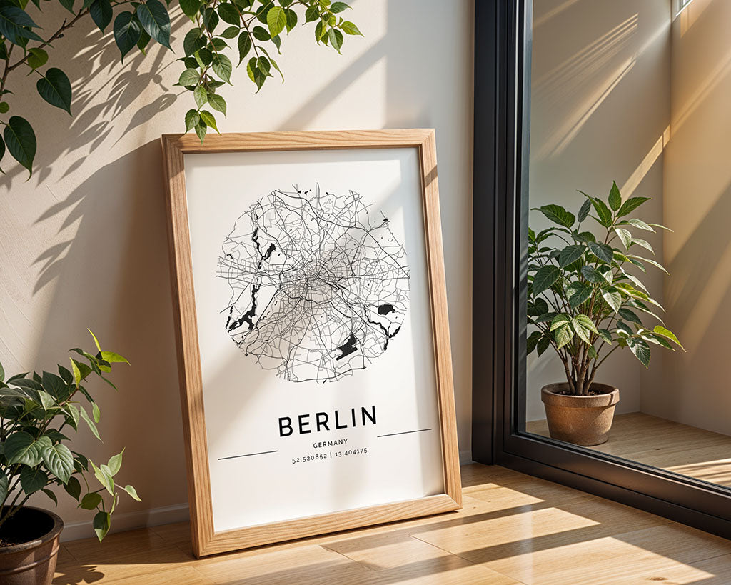 A framed FN Prints Berlin City Map rests on a sunlit wooden floor near a glass door. This minimalist poster showcases the city's coordinates, with potted plants beside it and leafy vines hanging above, creating a cozy indoor setting.