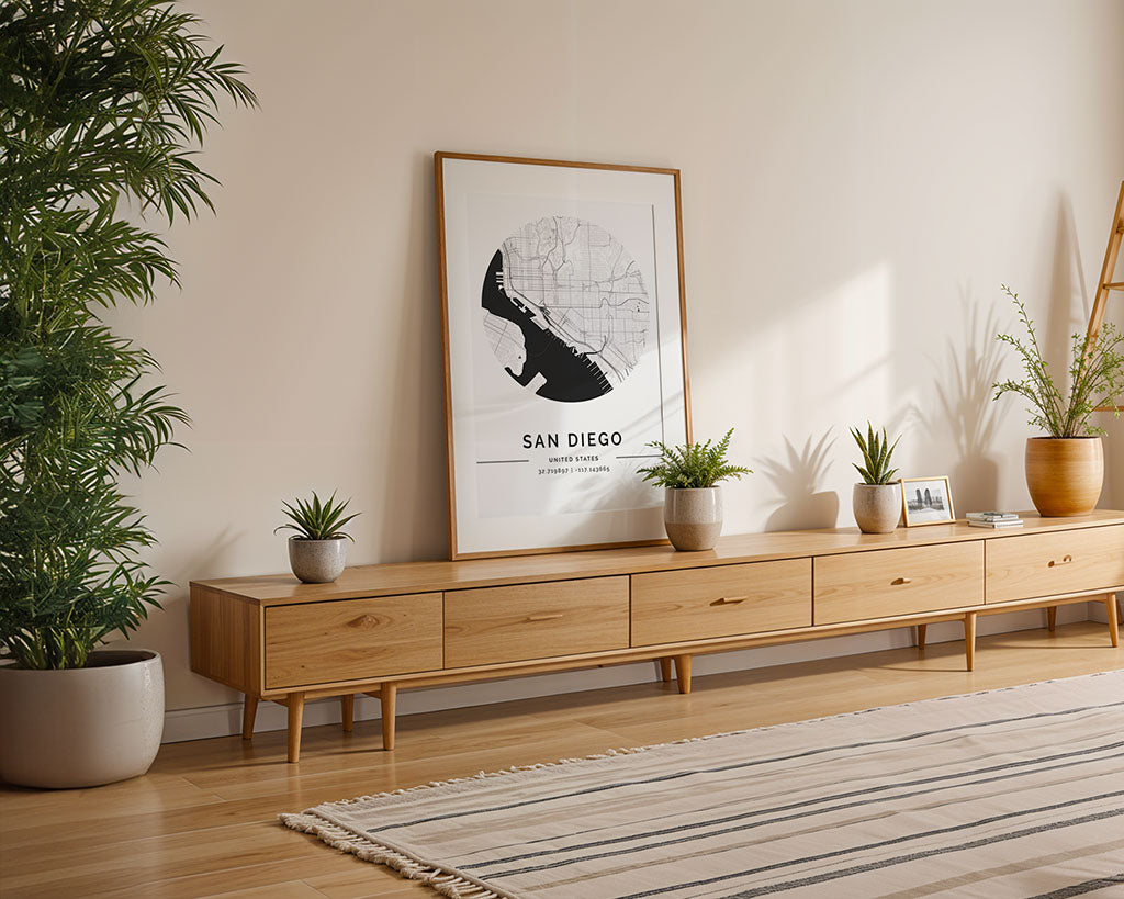 A light wood sideboard with potted plants sits against a beige wall. The FN Prints San Diego City Map rests on top, adding minimalist flair. A striped rug covers the hardwood floor, with a large plant on the left.