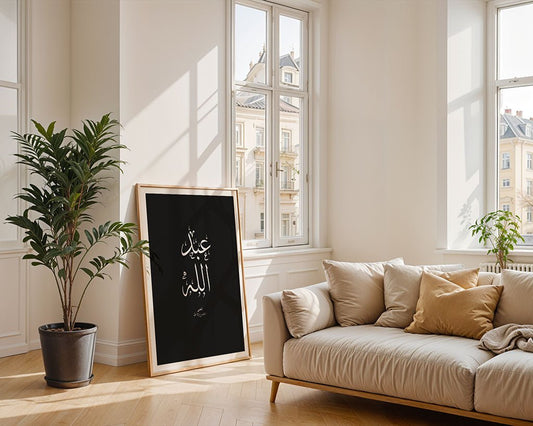 The "Abdulla - عبدالله" framed Arabic calligraphy wall art by FN Prints rests beside a beige sofa in a bright living room, showcasing Islamic tradition.