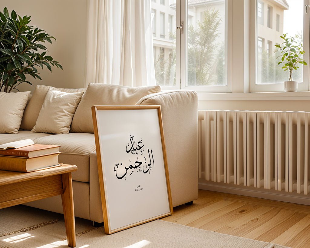 The Abdulrahman - عبدالرحمن framed Islamic wall art by FN Prints leans against a beige sofa in a sunlit living room, with books stacked on the armrest, a potted plant on the windowsill, and sunlight pouring through large windows.