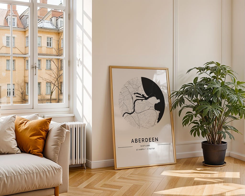A bright living room with a large window features a beige sofa with orange and white cushions, the FN Prints Aberdeen City Map leaning against the wall, a potted plant, and wooden herringbone flooring.
