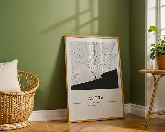 The Accra City Map by FN Prints, a minimalist framed poster of Ghana’s capital, is displayed on a wooden floor against a green wall beside a rattan chair and small table with a potted plant as sunlight casts gentle shadows.
