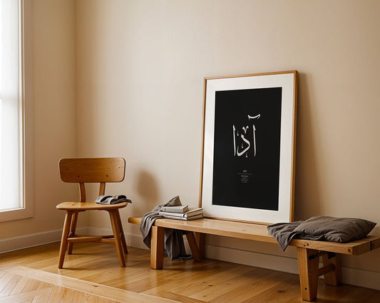 A minimal room features a wooden chair and bench with a cushion and books, plus "Ada - أدا" Arabic calligraphy wall art by FN Prints in a black frame, leaning against a beige wall on herringbone flooring. Natural light streams in from the left.