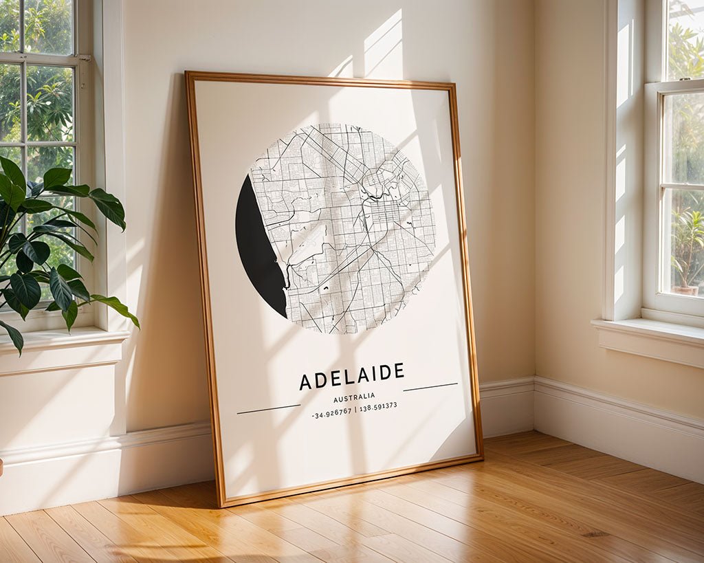 FN Prints' Adelaide City Map, a minimalist black and white circular design, is framed and placed against a wall in a sunlit room with wooden floors and large windows. A small plant beautifully complements this elegant piece of home décor.