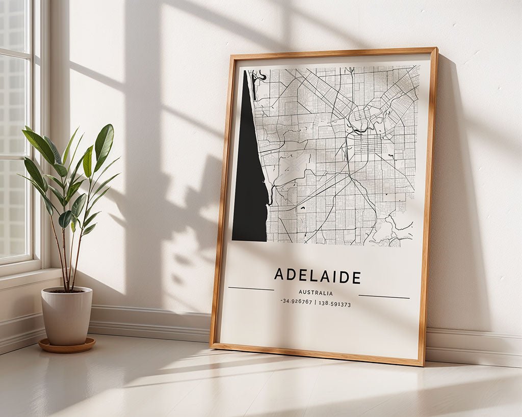 A framed FN Prints Adelaide City Map leans against a sunlit wall, reflecting minimalist design. Nearby, a potted plant with slender green leaves enhances the home décor as sunlight creates cozy shadows, fostering tranquility.