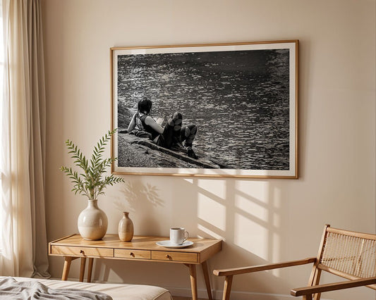 A wooden table with a vase, cup, and saucer sits beneath "Afternoon Read by the Canal," a black-and-white art photograph by FN Prints, hanging on a beige wall. A wooden chair is placed to the right of the table.
