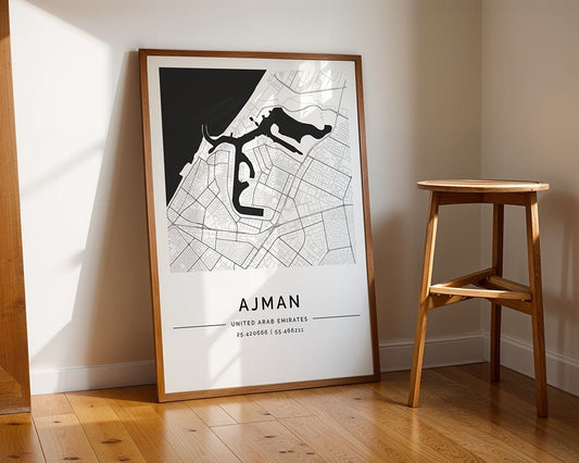 The Ajman City Map by FN Prints, a minimalist print on museum-quality fine art paper, rests against a white wall on a wooden floor beside a stool, with sunlight casting soft shadows for a tranquil display.