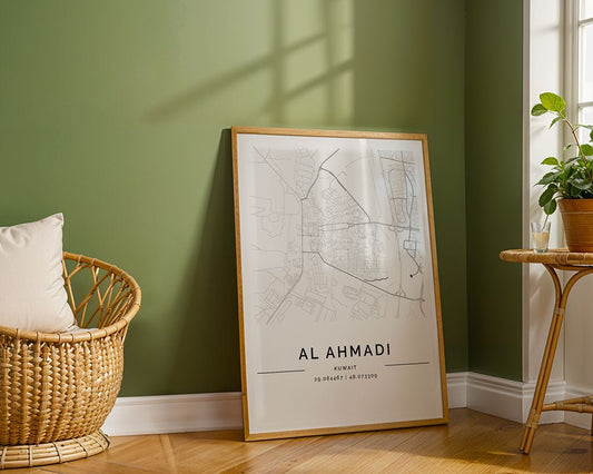 FN Prints Al Ahmadi City Map, a minimalist print on museum-quality fine art paper, leans against the green wall. A wicker chair with a white cushion is on the left and a potted plant on the right, as sunlight streams through the window, casting dancing shadows across the scene.