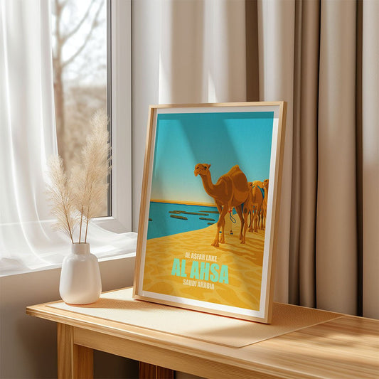 Framed artwork of camels on a beach with 'Al Ahsa' text, placed on a wooden surface near a window.