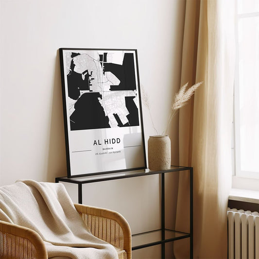 The FN Prints Al Hidd City Map minimalist framed print sits on a black metal console by a beige wall, next to a vase of dried grass and a draped blanket on a wicker chair, with sunlight streaming through the window.