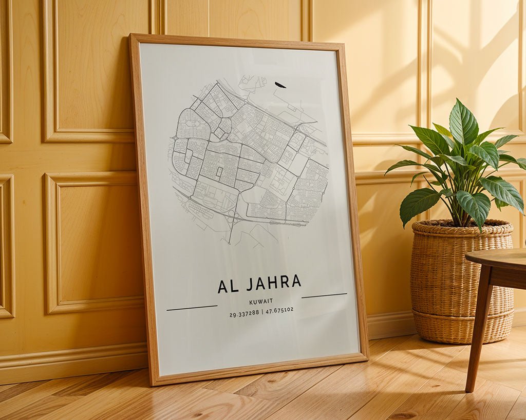 An FN Prints "Al Jahra City Map" framed minimalist print on fine art paper leans against a light yellow paneled wall, showcasing geographical coordinates. To its right, a green potted plant in a wicker basket rests on the wooden floor.