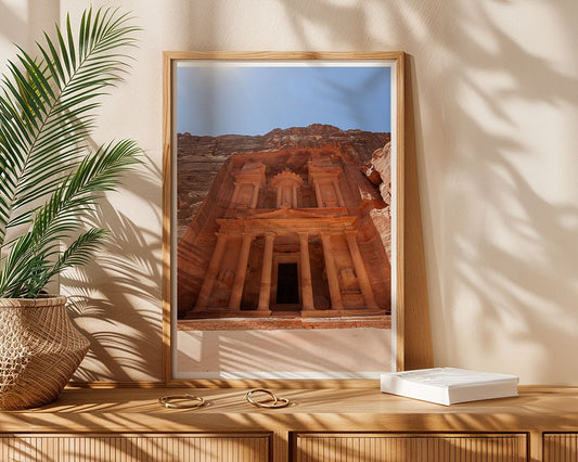 A framed FN Prints "Al-Khazneh - The Treasury - Petra: Part II" photograph sits on a wooden console table next to a green potted plant and closed white book, dappled with sunlight and shadow.