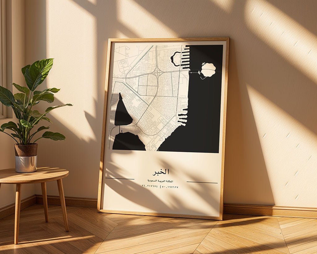 The FN Prints Al Khobar City Map, framed and leaning against a wall on a wooden floor, is illuminated by sunlight beside a round table with a green potted plant—a minimalist Saudi Arabia-inspired décor piece.