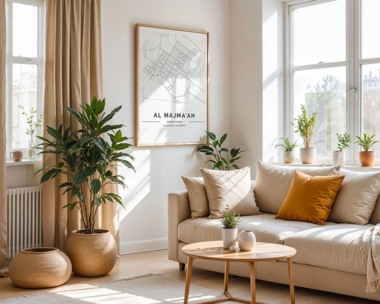 A bright living room with large windows, beige curtains, a light sofa with yellow and beige pillows, potted plants, a round wooden coffee table, and the FN Prints Al Majma'ah City Map displayed on the wall.