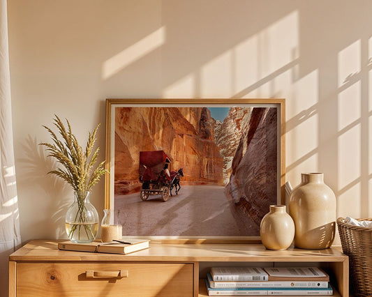 The "Al Siq Petra" framed photo by FN Prints rests on a wooden cabinet beside beige ceramic vases, a glass vase with dried grass, and stacked books in a sunlit room.