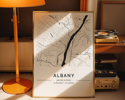 A framed Albany City Map by FN Prints rests against the wall in a cozy room. Printed on museum-quality paper, it accompanies a vintage lamp and record player on a wooden table. The bottom of this city wall art displays Albany's coordinates: 42.652628, -73.756409.