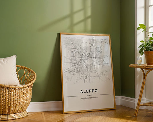 An FN Prints Aleppo City Map is framed and leans against a green wall, displaying detailed street layouts. A wicker chair and table with a potted plant enhance the Scandinavian décor, while sunlight filters in, casting delightful shadows.