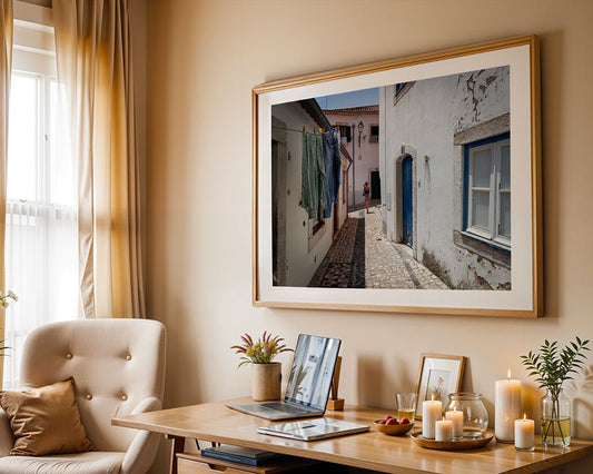 A home office with a beige chair, wooden desk, candles, and laptop is completed by FN Prints’ "Alfama Explorer" above the desk—serene wall art depicting Alfama’s cobblestone alley, hanging laundry, and white buildings.