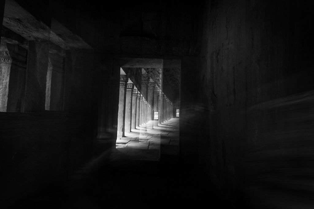 Dramatic black and white fine art photography of a stone corridor in an ancient Cambodian temple with light rays streaming through pillars.