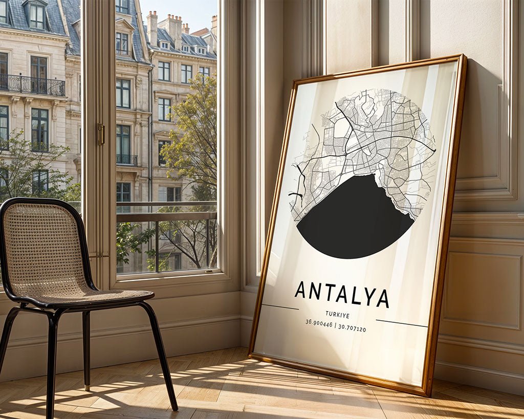 An Antalya City Map by FN Prints, framed and crafted on museum-quality art paper, leans against a sunlit room's wall, showcasing its circular design and coordinates. A woven chair complements the scene as large windows display an urban view of trees and historic buildings.