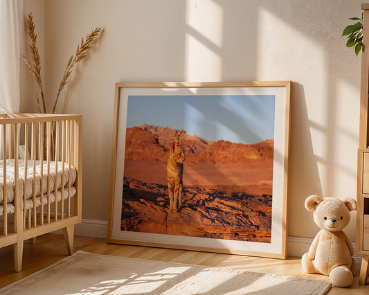 The framed "Antar, The Wadi Rum Cat, Jordan" art print by FN Prints depicting a cheetah rests on the nursery floor beside a wooden crib and beige teddy bear. Dried grasses in a vase add warmth to the cozy space.