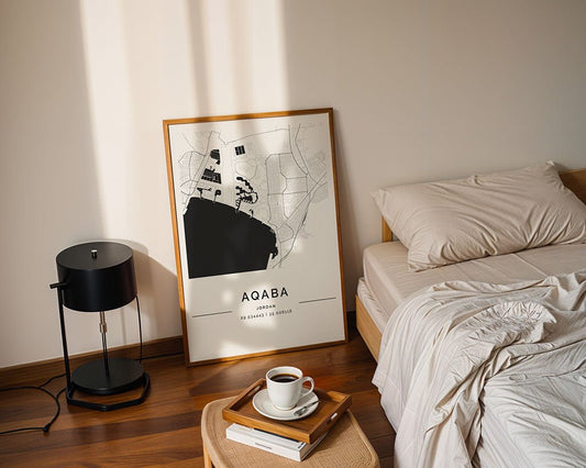 In a sunlit cozy bedroom, beige linens dress the bed next to a round black lamp on a table. A FN Prints' Aqaba City Map rests framed against the wall. On the bed, a wooden tray with a coffee cup and book offers relaxation.