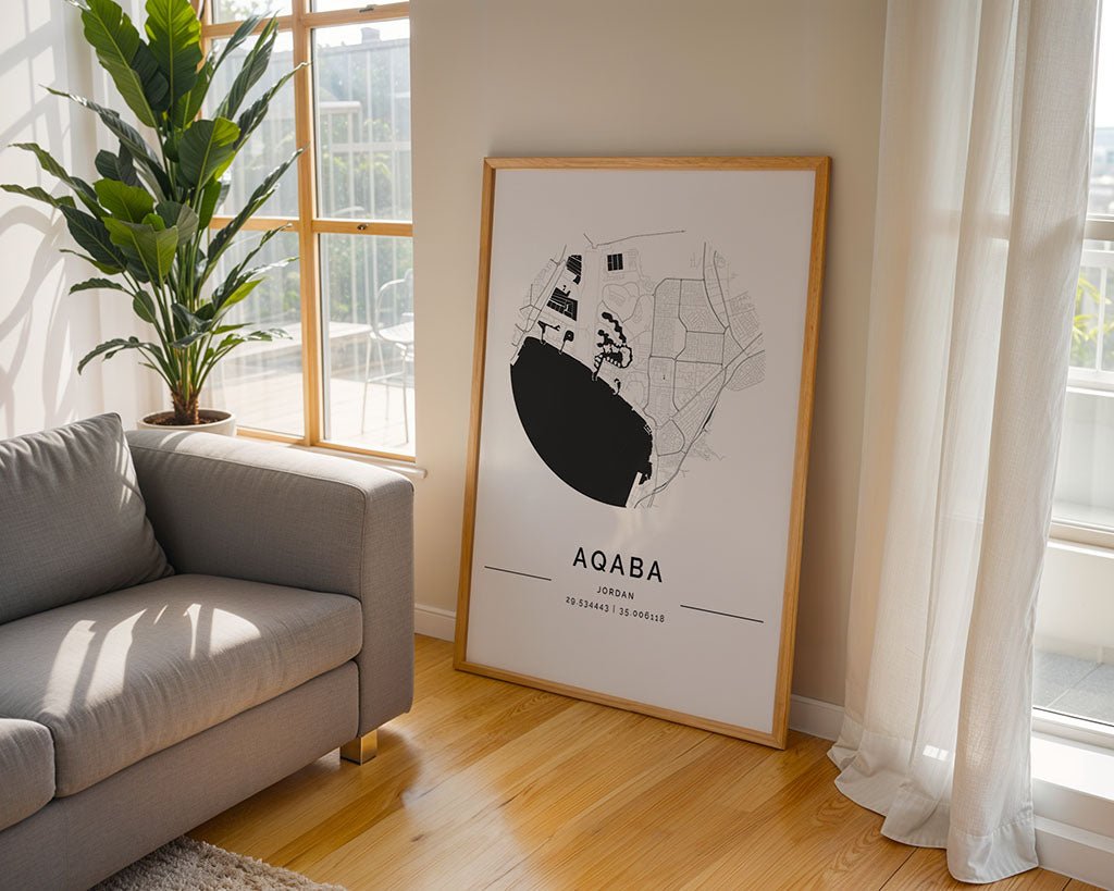 The FN Prints' Aqaba City Map, a large framed minimalist artwork, is propped against a light wall in a bright room. Nearby is a gray sofa with sheer-curtained windows and a tall green plant, all crafted through a sustainable printing process.
