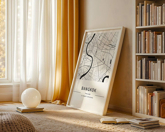 A framed FN Prints Bangkok City Map leans against a wall in a cozy room with light curtains and a sunlit window, featuring a neatly arranged bookshelf. Urban design fans will love the small globe light on the carpeted floor next to stacked books.