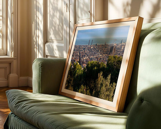 The framed "Barcelona II" cityscape by FN Prints sits on a green sofa in a sunlit room, displaying Barcelona's buildings, trees, and ocean beneath a clear sky as window shadows softly cross the scene.