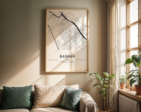 FN Prints' framed Basra City Map on museum-quality paper hangs above a beige sofa with green and beige cushions. Sunlight through the window highlights a small plant on a side table, adding to the room's cozy ambiance.