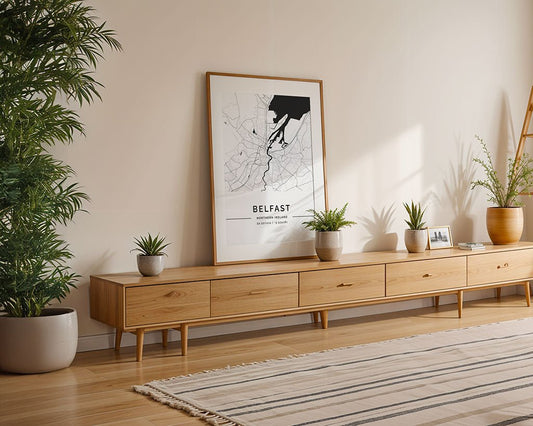 A minimalist living room features a sleek, wooden sideboard with drawers. A FN Prints Belfast City Map leans against the wall, printed on museum-quality fine art paper. Potted plants decorate the sideboard and floor, while a striped rug enhances the wood floor under a ladder shelf.