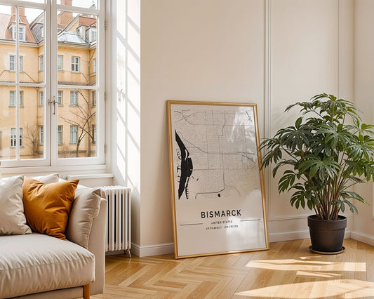 A sunlit room features a beige sofa with orange and white cushions near a large window. A potted plant and FN Prints' Bismarck City Map decorate the wooden herringbone floor, adding elegance. An apartment building is visible outside.