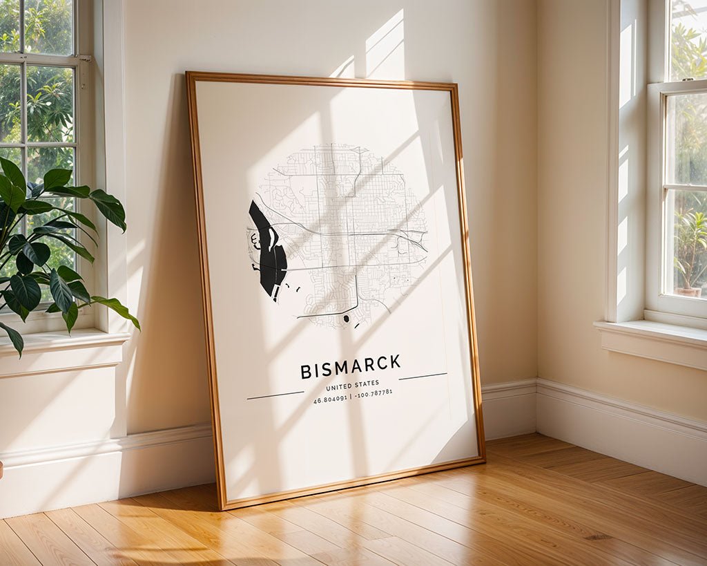 A framed FN Prints' Bismarck City Map leans in a sunlit room with sunlight casting shadows through a window, and a leafy green plant rests on the wooden floor beside the frame.