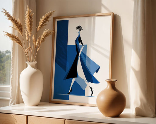 The Blue Minimalist Woman framed wall art by FN Prints stands on a light wood surface beside two neutral vases with dried grasses, illuminated by sunlight from a nearby window.