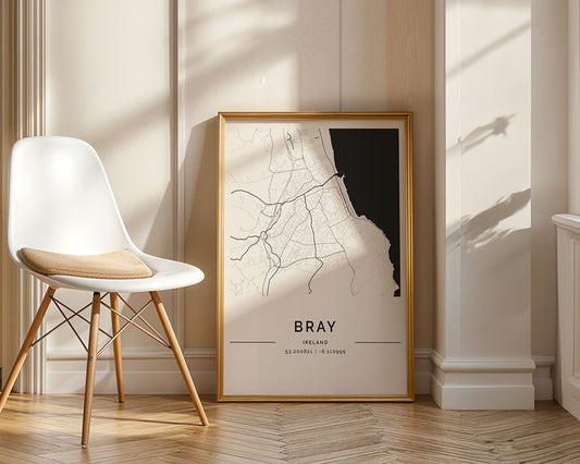 A framed FN Prints Bray City Map, a minimalist museum-quality art piece, leans against a sunlit wall as shadows gently dance. Nearby, a cushioned white chair with wooden legs awaits.