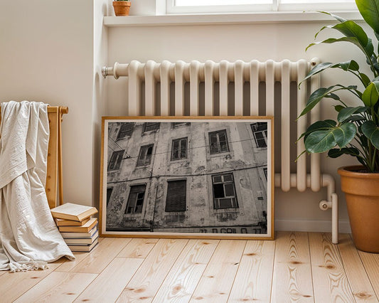 The "Broken Windows Black & White" framed urban photograph by FN Prints leans against a radiator on a light wood floor, complemented by a folded blanket, books, and a potted plant for a charming vintage architectural display.