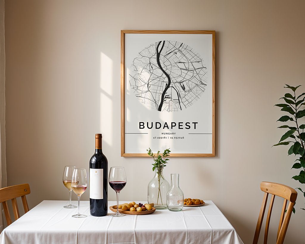 A dining table with a white cloth is elegantly set with red wine, two glasses, a carafe, and snacks. Above hangs the FN Prints Budapest City Map in a wooden frame. A small vase of flowers adds charm to the scene.