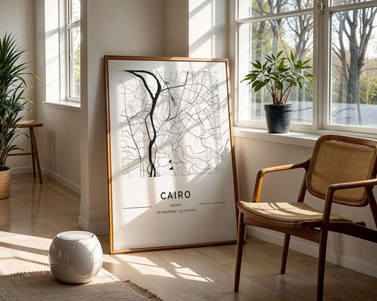 An FN Prints Cairo City Map, printed sustainably and framed, leans against a sunlit wall. A chair and stool enhance the room's decor, complemented by indoor greenery and a view of trees outside. The wooden floor adds warmth to this serene setting.