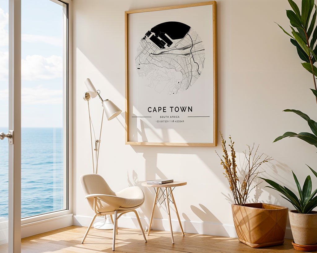 A minimalist room features a FN Prints "Cape Town City - Map" on the wall. Nearby, a white modern chair and a small wooden table are illuminated by a floor lamp. A large window reveals ocean views, while greenery from a plant enhances the tranquil ambience.