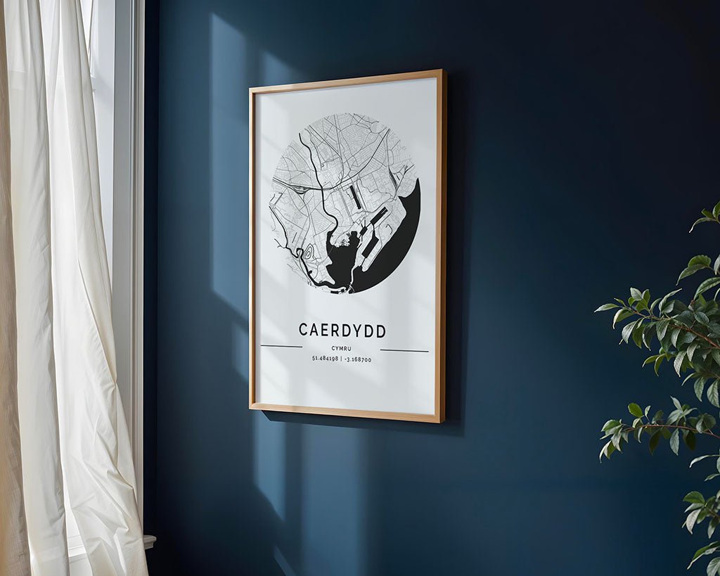 The Cardiff City Map by FN Prints is displayed on a dark blue wall next to sheer-curtained window. A green plant peeks from the corner, casting playful shadows on the museum-quality fine art paper print.