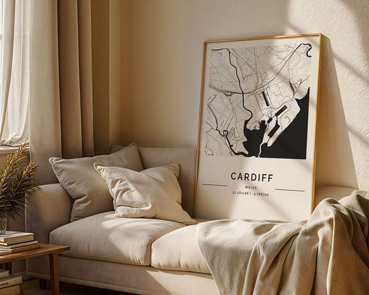 In a sunlit room, the FN Prints Cardiff City Map leans elegantly against a beige couch. A cushioned beige throw is draped across it, and nearby, a stack of books enhances the serene setting.