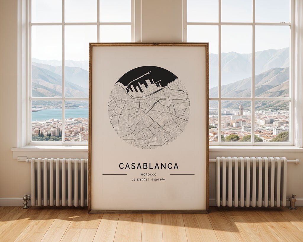 A large framed FN Prints Casablanca City Map leans against a bright room's wall, elegantly capturing the city's circular streets and coordinates. Ideal as a minimalist print or thoughtful housewarming gift, large windows reveal a stunning cityscape and distant mountains.