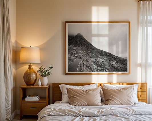 A neatly made bed with beige bedding stands against the wall. Above it hangs "Connor Pass Black & White" by FN Prints. A wooden nightstand holds a lamp, a plant, and two white vases as sunlight streams through a nearby window.