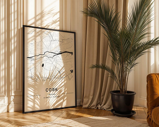 A black-framed, eco-friendly Cork City Map by FN Prints leans against a cream wall with long curtains. Sunlight casts grid shadows on the minimalist print. To the right, a potted palm complements an orange sofa slightly visible in the scene.