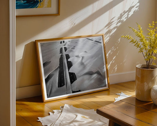 Desert Roads by FN Prints, a framed black and white desert photograph featuring a road through sand dunes, leans against the wall on a wooden floor near a small table with a potted plant. Sunlight and shadows highlight this minimalistic scene.