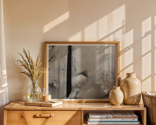 A wooden cabinet displays FN Prints' Desert Roads IV framed artwork, two beige vases, a glass vase of dried wheat, and stacked books—modern decor illuminated by sunlight and plant shadows on the wall and cabinet.